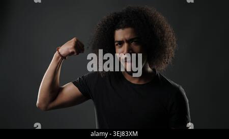 Jeune homme debout, regardant la caméra, fléchissant le biceps montrant la force et la confiance Banque D'Images