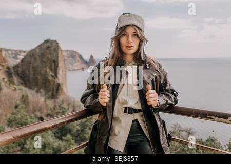 Femme voyage côte jeune voyageuse femme portant chapeau ushanka et veste en cuir posant contre les falaises de mer rocheuses en automne Banque D'Images
