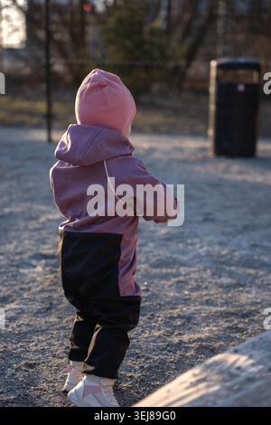 un tout-petit rétro-éclairé dans un bonnet rose et un vêtement d'extérieur violet debout sur du gravier dans un parc de jeux urbain calme pendant une heure d'or fraîche, un bokeh doux Banque D'Images