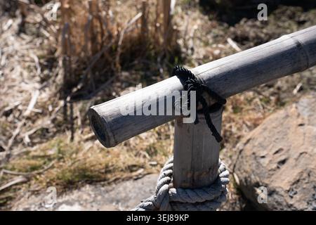 poteau en bambou vieilli fixé à un poteau en bois avec une corde noire enroulée et une épaisse ligne de fibres naturelles, reposant sur une banque rocheuse ensoleillée au milieu d'herbes sèches Banque D'Images