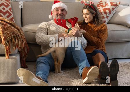 Couple assis sur un tapis en peluche dans le salon caressant chiot portant des bois rouges et chapeau de Père Noël. Confortable, maison, duo, chien, festif, lumière du jour, décor Banque D'Images