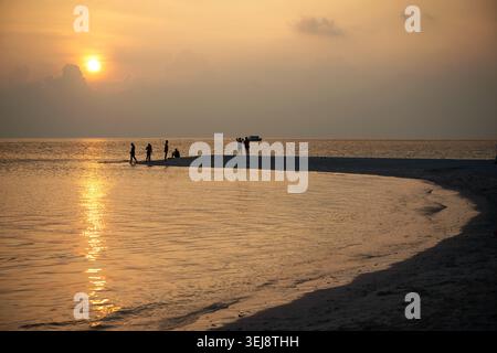Une photographie grand angle capturant un groupe de personnes silhouettées contre un coucher de soleil vibrant sur un banc de sable étroit. Le soleil bas projette une lueur dorée chaude acr Banque D'Images