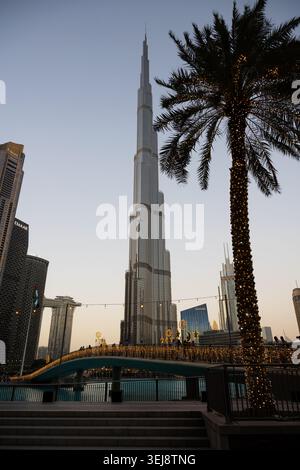 Une photographie verticale en bas angle du gratte-ciel Burj Khalifa à Dubaï au crépuscule. Au premier plan, un grand palmier est enveloppé de déc. Lumineux Banque D'Images
