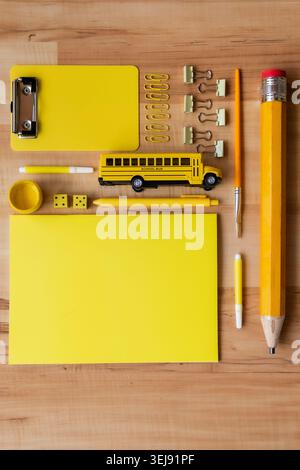 Pose à plat créative de fournitures scolaires jaunes, y compris un crayon géant, un autobus scolaire jouet et des blocs-notes soigneusement disposés sur un bureau en bois. Retour à l'école Banque D'Images