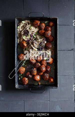 op view de nouilles soba servies avec des boulettes de viande glacées brillantes dans un plateau rustique en métal noir. Les nouilles sont garnies de rondelles d'oignons verts et de purp Banque D'Images