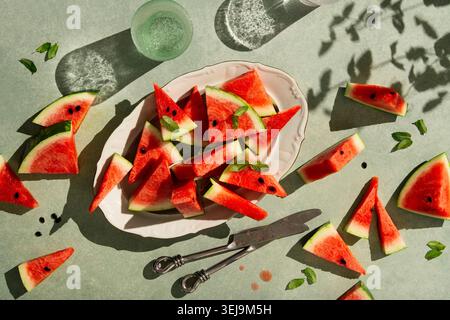 pose à plat de pastèque tranchée dans une assiette ovale stylisée avec des feuilles de menthe fraîche et deux verres d'eau pétillante en haute clé Banque D'Images