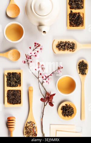 Pose plate minimale de théière, tasses à thé, cuillères en bois avec des variétés de thé en vrac et branche de fleur de cerisier sur un fond clair. Préparation de thé élégante Banque D'Images