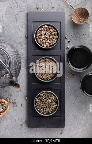 Pose plate de variétés de thé en vrac assorties dans des bols avec théière en fonte, tasses et passoire sur un fond de pierre sombre. Dégustation de thé asiatique élégante comp Banque D'Images