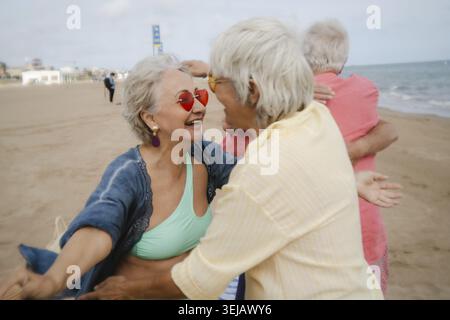 Femmes âgées heureuses embrassant sur une plage de sable, riant tout en portant des lunettes de soleil en forme de cœur, avec une troisième personne serrée dans les bras en arrière-plan, incardyin Banque D'Images