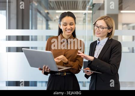Deux femmes d'affaires diverses discutant de stratégie de projet à l'aide d'un ordinateur portable et d'une tablette tout en se tenant debout dans un cadre de bureau contemporain, représentant le travail d'équipe et la communication professionnelle Banque D'Images