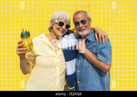 Couple senior souriant et embrassant contre un mur carrelé jaune vibrant, portant des lunettes de soleil, femme tient la boisson à l'ananas, rayonnant de joie de vacances, Activ Banque D'Images