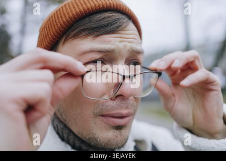 Un jeune homme portant un bonnet orange et une veste polaire blanche ajuste ses lunettes tout en éprouvant une irritation oculaire dans un cadre extérieur Banque D'Images