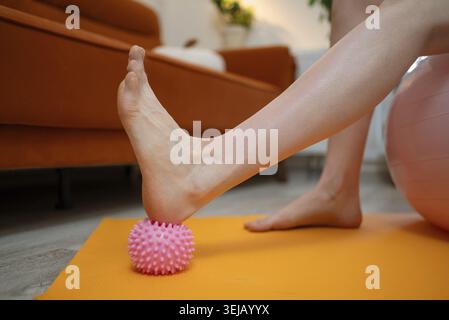 Femme enceinte renforçant et étirant ses pieds à l'aide d'une boule de massage rose sur un tapis de yoga orange vif à l'intérieur, en donnant la priorité à l'auto-soin et au séjour Banque D'Images