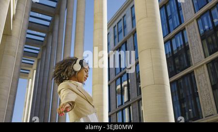 Avec des écouteurs et un sourire radieux, une jeune femme professionnelle danse librement dans un espace urbain, son mouvement dynamique contrastant avec le l statique Banque D'Images