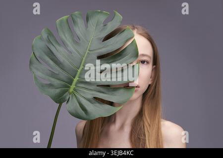 Jeune modèle féminin caucasien avec la feuille de monstera couvrant le visage sur fond gris Banque D'Images