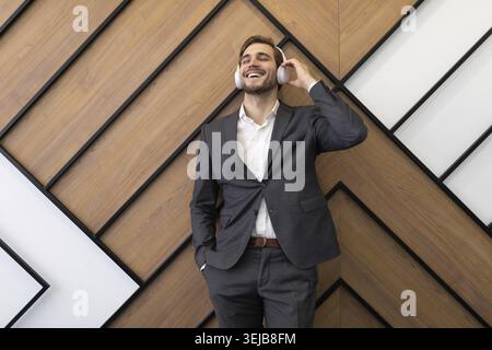 Un jeune homme d'affaires dans un costume gris strict aime écouter de la musique sur des écouteurs sur le fond d'un mur de bureau Banque D'Images