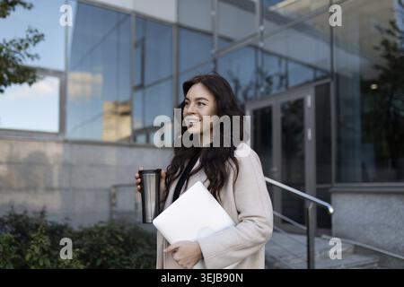 Confiante femme d'affaires moderne va travailler avec un grand sourire avec un ordinateur portable dans ses mains Banque D'Images