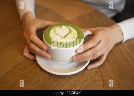 Mains de femme tenant une tasse blanche de latte matcha décorée d'un art latte en forme de cœur, placée sur une table en bois, créant un atmo confortable et invitant Banque D'Images