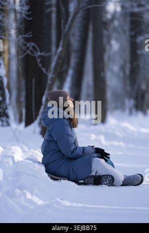 Jeune femme vêtue d'une tenue d'hiver confortable, assise sur un traîneau dans la forêt enneigée, profitant de la chaleur du soleil d'hiver tout en embrassant la tranquillité Banque D'Images