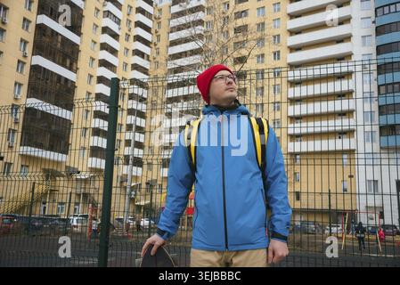 Livreur de nourriture avec sac à dos jaune et veste bleue tenant skateboard et regardant devant les bâtiments résidentiels, portant des lunettes et rouge Banque D'Images