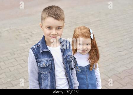 Enfant masculin caucasien en veste denim avec enfant femelle aux cheveux rouges souriant à l'extérieur sur une surface pavée Banque D'Images