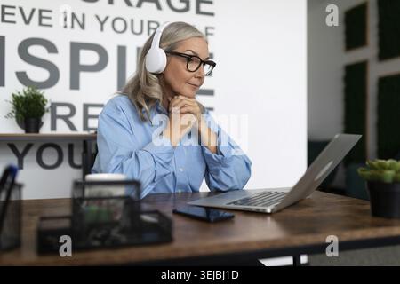 Entreprise sérieuse femme d'affaires mature regardant un séminaire en ligne sur un ordinateur portable au bureau avec ses mains repliées sous son menton dans un casque Banque D'Images