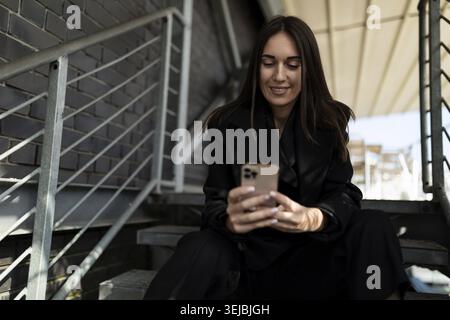 Femme d'affaires mature dans un costume d'affaires avec un téléphone portable dans ses mains avec un sourire se trouve sur les marches à côté du bureau Banque D'Images