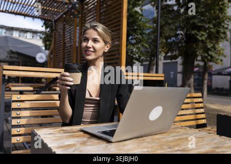 Femme d'affaires élégante buvant du café et travaillant sur la terrasse avec ordinateur portable à l'extérieur Banque D'Images