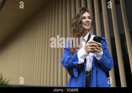 Jeune étudiante avec un téléphone portable regardant au loin avec un sourire sur fond de bandes verticales dorées Banque D'Images