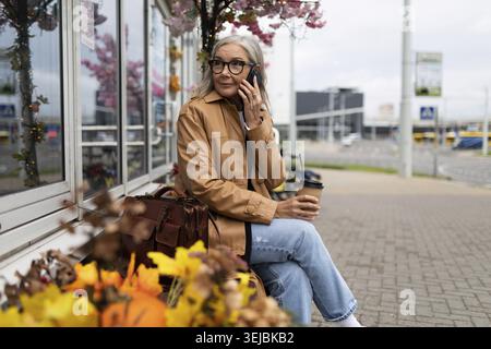 Femme âgée parlant au téléphone assise sur un banc près du magasin Banque D'Images