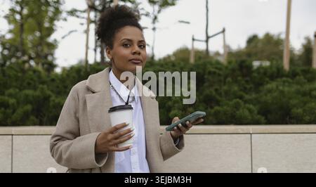 Femme d'affaires afro-américaine élégante avec une tasse de café et un téléphone dans ses mains à l'extérieur Banque D'Images