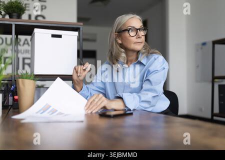 Âge femme caucasienne homme d'affaires dans des lunettes avec des documents est assis au bureau Banque D'Images