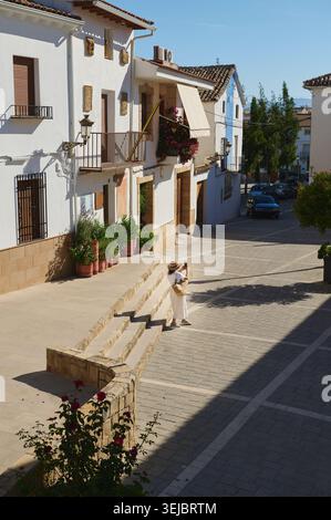 Femme voyageant à travers une rue de village espagnol ensoleillée avec des maisons blanchies à la chaux, des balcons, des fleurs en pot et des montagnes lointaines, profitant de la détente Banque D'Images
