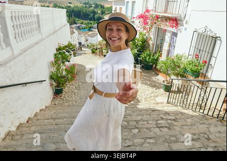 Femme souriante voyageant à travers un village espagnol blanchi à la chaux dans un chapeau de soleil et une jupe, menant un compagnon par la main sur les marches pavées ensoleillées surr Banque D'Images