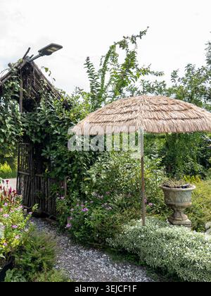 Jardin tropical rustique avec une arche de bambou, une végétation luxuriante, des plantes à fleurs, un chemin de gravier et un parasol de chaume dans un paisible pays en plein air Banque D'Images