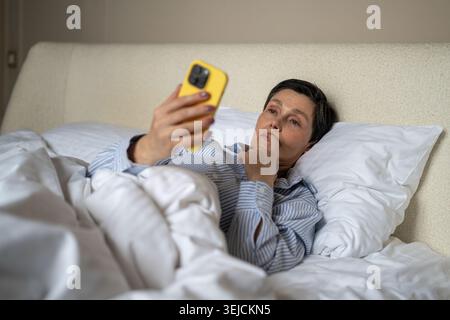 Femme âgée réfléchie dans le lit procrastinant regarder les nouvelles en ligne sur smartphone après le réveil. Banque D'Images