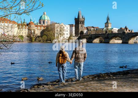 Un couple explore le charmant front de mer de Prague, main dans la main, tandis que les canards nagent. L'architecture emblématique et le ciel bleu vif créent un cadre pittoresque pour leur après-midi mémorable. Banque D'Images