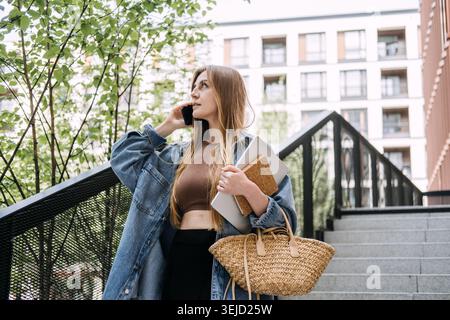 Jeune femme parle sur smartphone tout en descendant les escaliers extérieurs de la ville tenant un ordinateur portable. Exécution agile des flux de travail, activité rapide, indépendant Banque D'Images