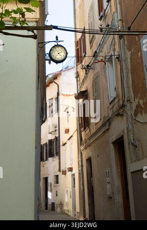 Piran, Slovénie. 26 mai 2026. Une horloge vintage est suspendue au-dessus d'une rue étroite bordée de vieux bâtiments. La lumière du soleil illumine les façades et les moulages vieillissants Banque D'Images