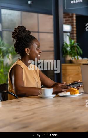Femme africaine adulte travaillant à la table de café en utilisant le téléphone et l'ordinateur portable, portant le dessus beige, boucles d'oreilles cerceau Banque D'Images