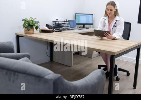 Médecin adulte de sexe féminin portant une blouse de laboratoire, gommages roses assis bureau clinique examinant la tablette, espace de copie Banque D'Images