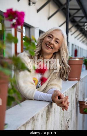 Femme mature souriante appuyée sur le balcon avec des fleurs roses Banque D'Images