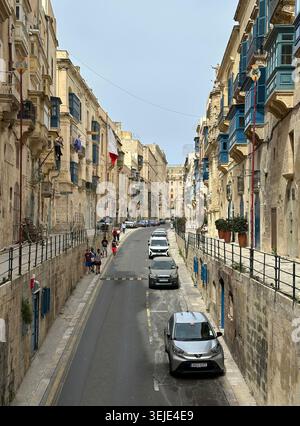 Rue étroite de la Valette, Malte, bordée de bâtiments historiques en calcaire avec balcons en bois coloré. Plusieurs véhicules sont stationnés le long de la R. Banque D'Images