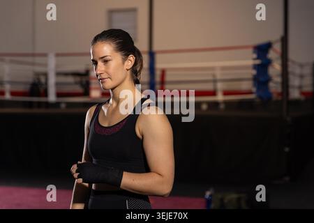 Boxeur féminin portant un débardeur noir ajustant les mains enveloppées tout en se tenant près de l'anneau de boxe dans le gymnase. Athlète, entraînement, enveloppement, concentration, discipline, cordes, arène Banque D'Images