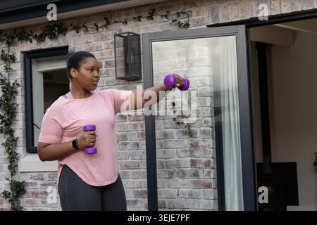 Femme afro-américaine exerçant sur le patio tenant des haltères violets en tee-shirt rose montrant une montre noire. Poids, séance d'entraînement, terrasse, jardin, Candid, fitness, bien-être Banque D'Images
