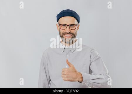 Homme souriant positif avec chaume, lève le pouce vers le haut, démontre son amour et son approbation, porte un couvre-chef et une chemise formelle, isolé sur fond blanc. Ma réponse oui. Gestes. Langage corporel Banque D'Images