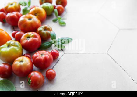 Tomates héritées fraîchement récoltées dispersées sur une surface carrelée hexagonale beige avec des feuilles de basilic Banque D'Images
