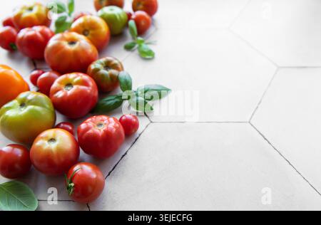 Tomates héritées fraîchement récoltées dispersées sur une surface carrelée hexagonale beige avec des feuilles de basilic Banque D'Images