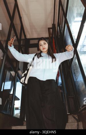 Confiant jeune femme asiatique dans la mode décontractée prend selfie avec smartphone tout en se tenant debout sur l'escalier. Cette photo en angle bas la montre regardant vers le bas avec l'esprit Banque D'Images