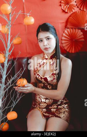 Élégante femme asiatique en robe qipao rouge moderne célèbre le nouvel an chinois. belle dame est assis tenant orange chanceux avec calme, expression séduisante Banque D'Images